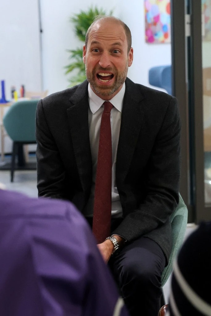 Prince William laughing heartily while seated and wearing a suit and red tie.